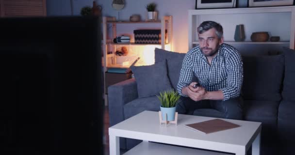 Medelålders kille tittar på TV hemma på natten på soffan ensam håller fjärrkontrollen — Stockvideo