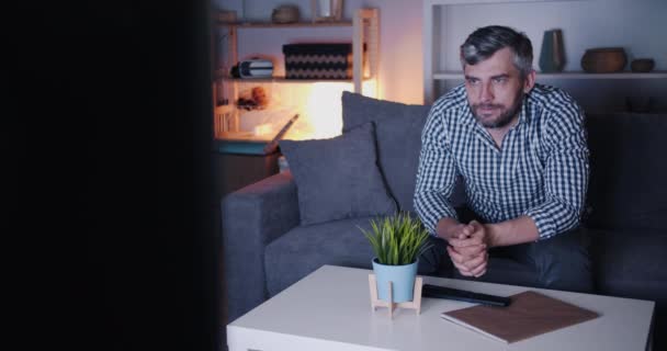 Hombre barbudo serio viendo la televisión por la noche en un apartamento oscuro sentado en un sofá — Vídeo de stock