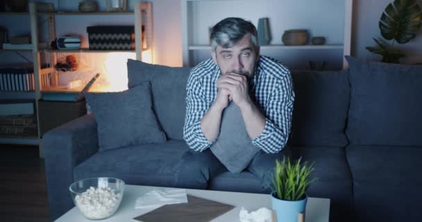 Портрет сумного хлопця, що витирає очі тканиною, дивлячись телевізор вдома вночі — стокове відео