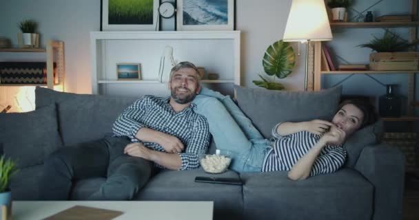 Joyful man and woman watcing TV throwing popcorn laughing in house at night — Stock Video