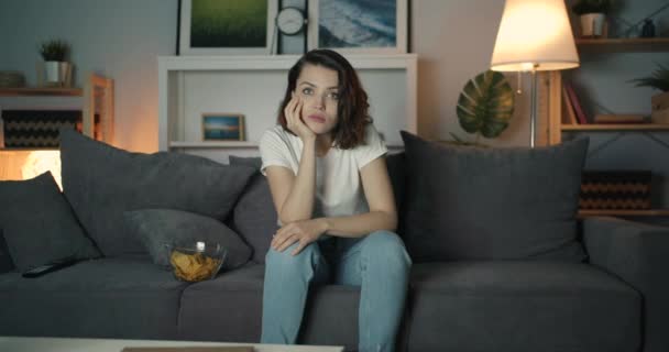 Young woman watching TV with serious face on couch at home at night — Stock Video