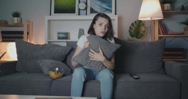 Young girl watching horror film on TV hiding behind pillow closing eyes at night — Stock Video