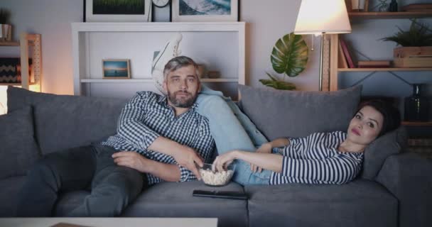 Chica y chico viendo la televisión por la noche comiendo palomitas de maíz en el sofá juntos — Vídeos de Stock