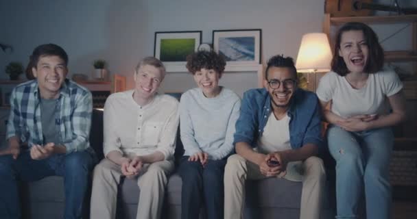 Amigos felizes assistindo TV rindo apontando para a câmera à noite em casa — Vídeo de Stock