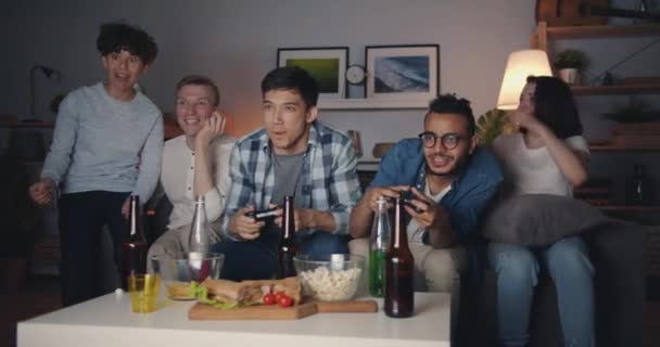 Багатоетнічна група друзів грають у відеоігри вдома пізно вночі розважаючись — стокове відео