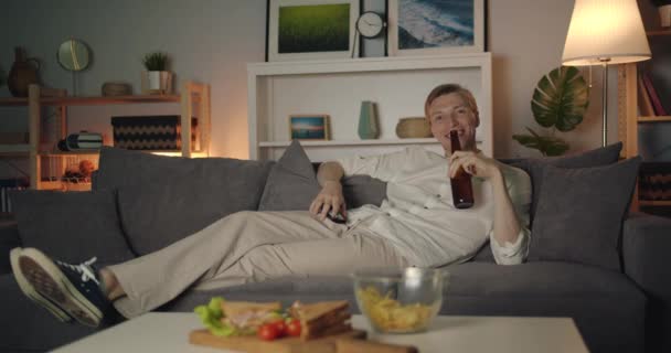 Fröhlicher Student, der Bier trinkt und allein in dunkler Wohnung vor dem Fernseher sitzt — Stockvideo