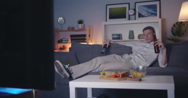 Joyeux jeune homme regardant la télévision boire de la bière se détendre sur le canapé la nuit — Video