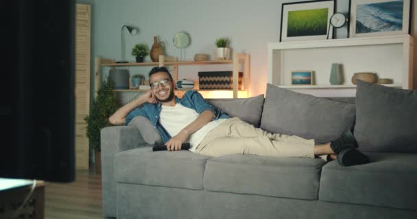 Беззаботный ближневосточный парень смотрит телевизор, смеясь, лежащий на диване в темном доме — стоковое видео
