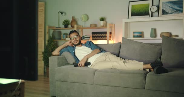 Уставший афроамериканец ночью смотрит телевизор и зевает на диване в одиночестве. — стоковое видео