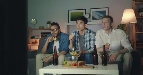 Vrienden kijken tv met aandacht eten snacks houden flessen 's nachts — Stockvideo