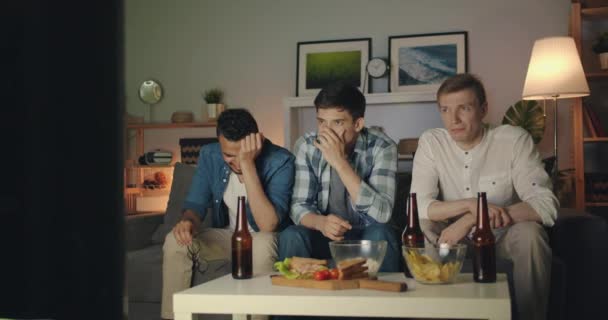 Hombres jóvenes infelices con caras tristes llorando viendo la televisión por la noche en el apartamento — Vídeos de Stock