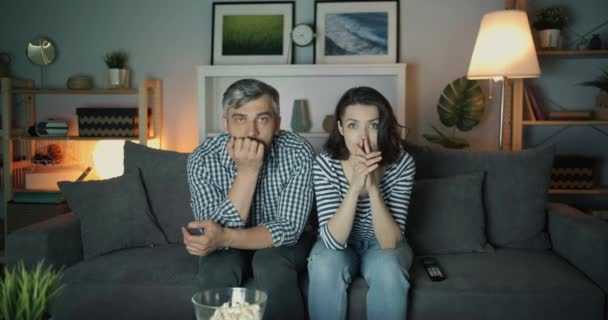 Шокированные девушка и парень смотрят телевизор с серьезными лицами трогательное лицо ночью — стоковое видео