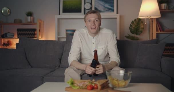 Портрет веселого парня, смотрящего телевизор, пьющего из бутылки, смеющегося ночью — стоковое видео