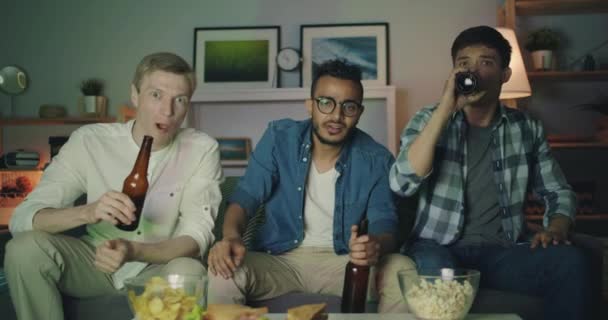 Guys watching sports on TV drinking eating snacks clinking bottles at night — Stock Video