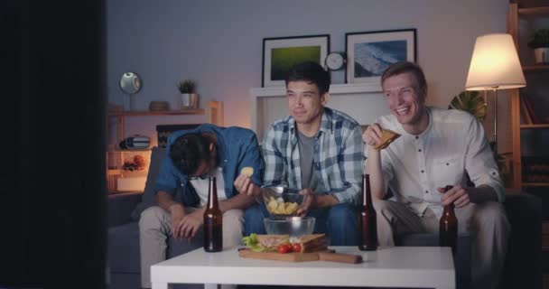 Χαρούμενοι τύποι που τρώνε σνακ βλέποντας αστεία show στην τηλεόραση γελώντας τη νύχτα στο σπίτι — Αρχείο Βίντεο