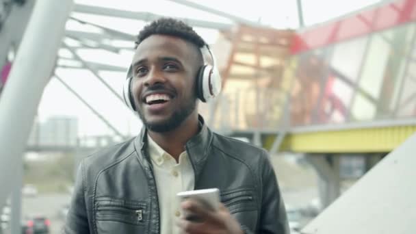 Estudiante en auriculares bailando al aire libre sosteniendo teléfono inteligente moderno — Vídeos de Stock