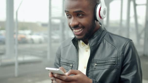 Knappe kerel in een koptelefoon met behulp van de smartphone lachend in City Street — Stockvideo