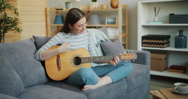 Hermosa mujer en gafas tocando la guitarra sentada en el sofá en casa en pose de loto — Vídeos de Stock