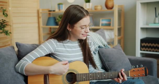 Девушка в очках играет на гитаре дома, наслаждаясь музыкой и отдыхом — стоковое видео