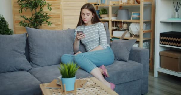 Αργή κίνηση της νεαρής κυρίας με σοβαρό πρόσωπο χρησιμοποιώντας smartphone στο σπίτι στον καναπέ — Αρχείο Βίντεο
