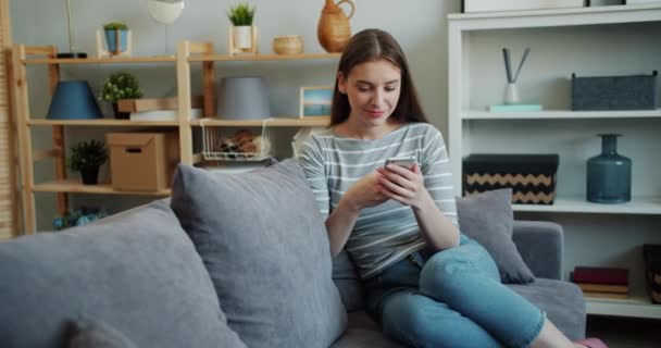 Chica bonita usando el teléfono inteligente moderno tocando la pantalla riendo en el sofá en casa — Vídeos de Stock