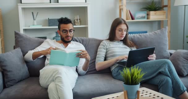 Дружина використовує ноутбук, коли чоловік читає книгу, сидячи на дивані вдома разом — стокове відео