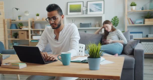 Hombre freelancer trabajando con el ordenador portátil en casa, mientras que la niña utilizando el teléfono inteligente en el sofá — Vídeos de Stock