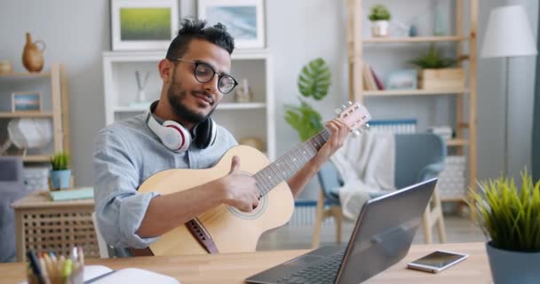 Joven árabe tocando la guitarra y usando el portátil en la mesa en el apartamento — Vídeo de stock
