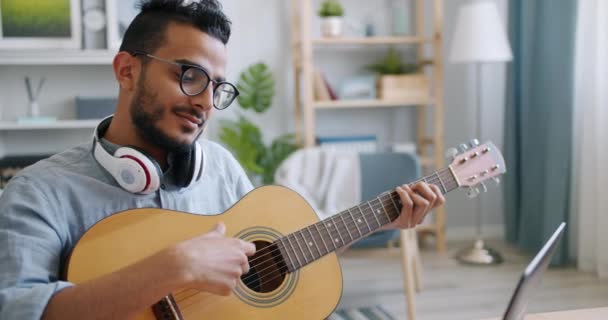 Cámara lenta del músico árabe tocando la guitarra frente a la computadora portátil en casa — Vídeos de Stock