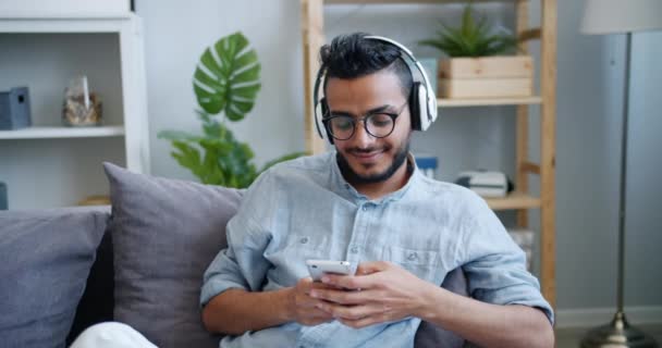 Pohledný arabský chlapík používající smartphone a poslech hudby ve sluchátkách doma — Stock video