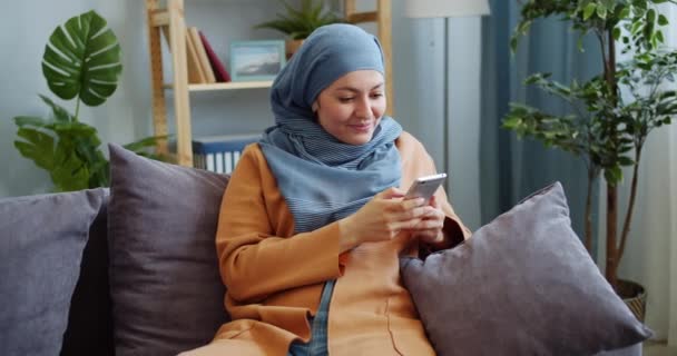 Menina alegre do Oriente Médio no hijab usando o smartphone rindo se divertindo em casa — Vídeo de Stock