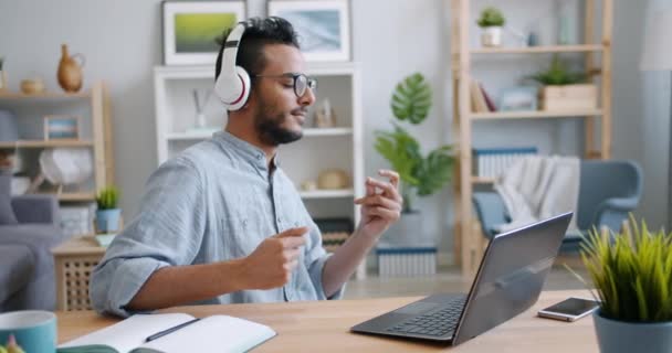 Beau jeune arabe appréciant la musique dans les écouteurs travaillant avec un ordinateur portable à la maison — Video