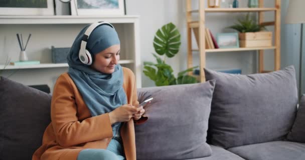 Chica musulmana alegre en hijab usando teléfono inteligente escuchando música en el apartamento — Vídeos de Stock