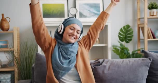 Cámara lenta de chica musulmana en hijab disfrutando de la música en los auriculares bailando en casa — Vídeos de Stock