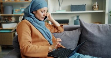 Hırslı Müslüman kız dizüstü bilgisayar ile çalışan ve evde cep telefonu konuşuyor