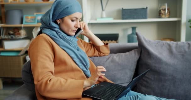 Chica musulmana ambiciosa trabajando con el ordenador portátil y hablando en el teléfono móvil en casa — Vídeos de Stock