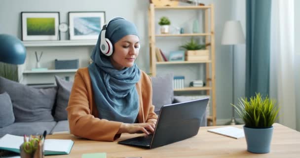 Müslüman kız dizüstü bilgisayar ile çalışan ve evde kulaklık müzik dinlerken — Stok video