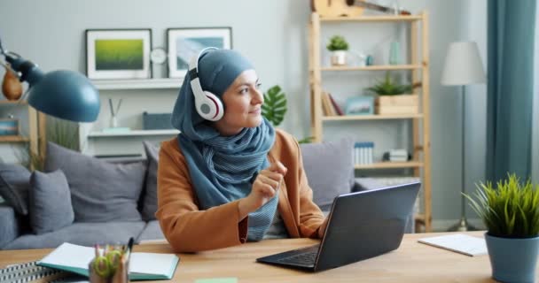 Blandad ras flicka i hijab njuter av musik i hörlurar med laptop hemma — Stockvideo