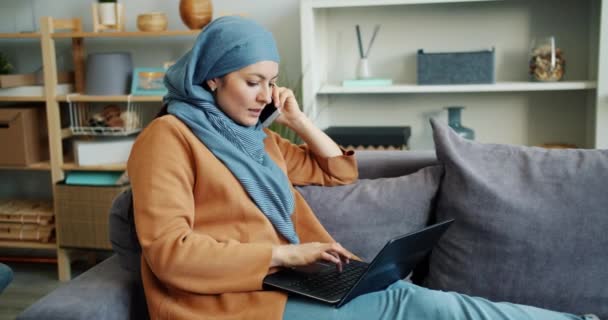 Cámara lenta de la mujer de negocios musulmana hablando en el teléfono móvil usando el ordenador portátil en casa — Vídeos de Stock