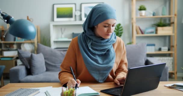 Chica musulmana freelancer trabajando con laptop y escribiendo en notebook en casa — Vídeos de Stock