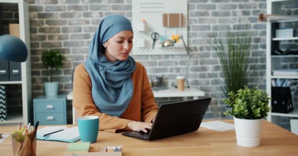 Mujer atractiva en hijab trabajando en la oficina con el ordenador portátil y luego mirando a la cámara — Vídeos de Stock