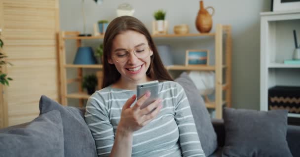 Привлекательный студент с помощью смартфона оставляет голосовое сообщение улыбаясь дома — стоковое видео