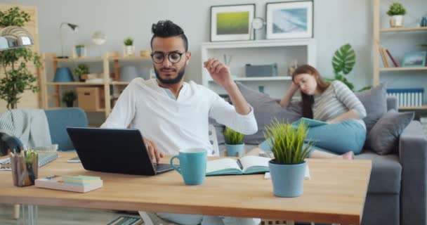 Уставший фрилансер, работающий с ноутбуком, расслабляется, пока женщина читает на заднем плане — стоковое видео