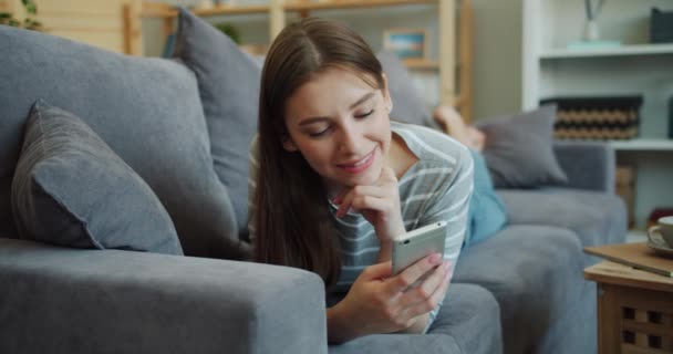 Bonita senhora usando smartphone e sorrindo deitado no sofá no apartamento — Vídeo de Stock