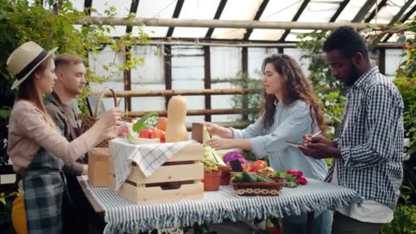 Pareja joven comprando alimentos orgánicos en el mercado agrícola en invernadero hablando con los agricultores — Vídeos de Stock