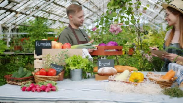 Mann bringt Gemüsekiste auf den Bauernmarkt und redet mit Verkäuferin im Gewächshaus — Stockvideo