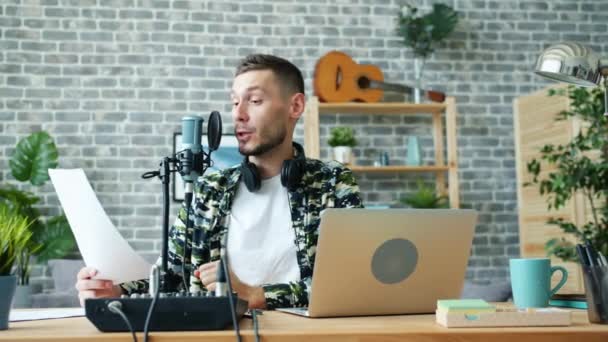 Jeune blogueur parlant au microphone en studio lisant du papier souriant — Video