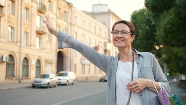 Весела дама з сумками махає рукою чекаючи таксі на відкритому повітрі в місті — стокове відео