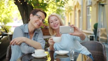 Sarışın akıllı telefon kullanarak kahve tutarak açık kafede arkadaşı ile selfie çekiyor