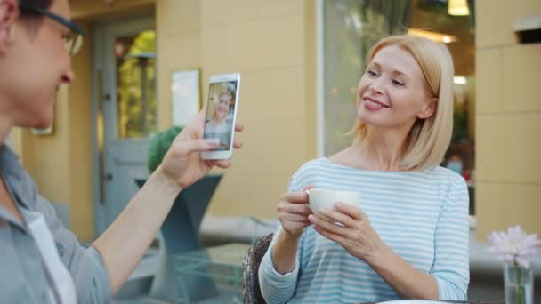 Amigo tomando fotos de la hermosa rubia madura sosteniendo taza de café en la cafetería de la calle — Vídeos de Stock